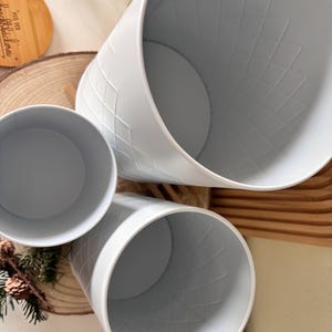 May include: Three light blue metal planters with a diamond pattern. The planters are cylindrical with a wide opening and a flat base. The planters are arranged at different angles, showcasing their interior and exterior details.