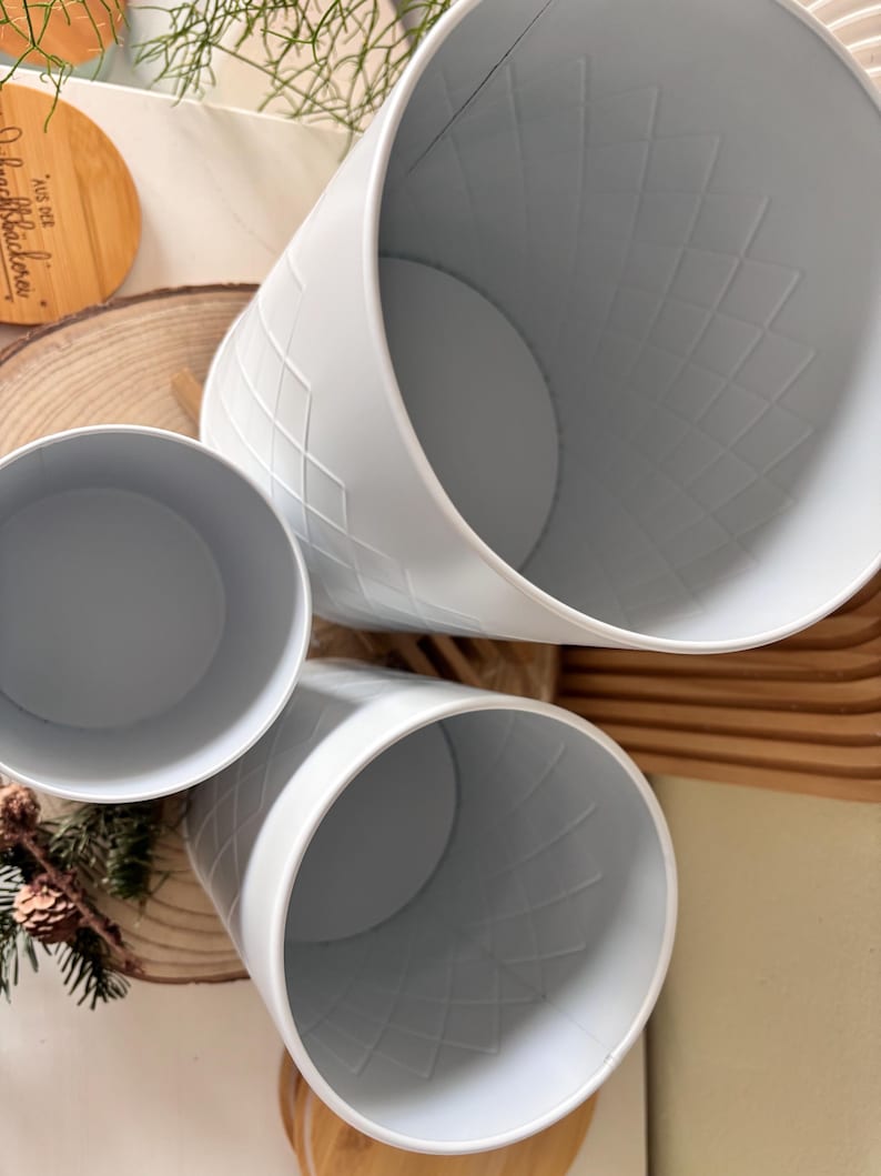 May include: Three light blue metal planters with a diamond pattern. The planters are cylindrical with a wide opening and a flat base. The planters are arranged at different angles, showcasing their interior and exterior details.