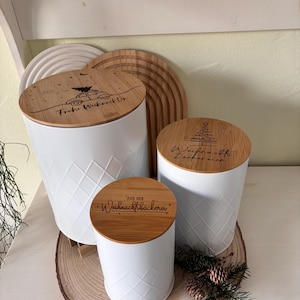 May include: Three white canisters with bamboo lids, featuring diamond patterns and engraved text. The lids have festive designs and phrases like "Frohe Weihnachten" and "Aus der Weihnachtsb&auml;ckerei". The canisters are on a wooden slice.