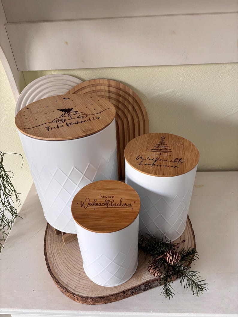 May include: Three white canisters with bamboo lids, featuring diamond patterns and engraved text. The lids have festive designs and phrases like "Frohe Weihnachten" and "Aus der Weihnachtsb&auml;ckerei". The canisters are on a wooden slice.