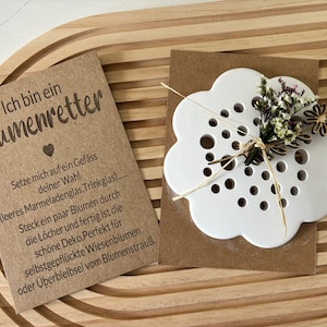 May include: A white ceramic flower-shaped vase with holes for stems. The vase is on a brown paper card with the text "Ich bin ein Blumenretter" and instructions for using the vase. The card and vase are on a wooden surface.