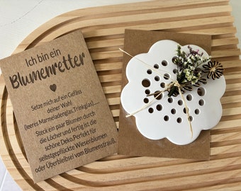 May include: A white ceramic flower shaped vase with holes for stems. The vase is on a brown paper card with the text "Ich bin ein Blumenretter" and instructions for using the vase. The card and vase are on a wooden surface.