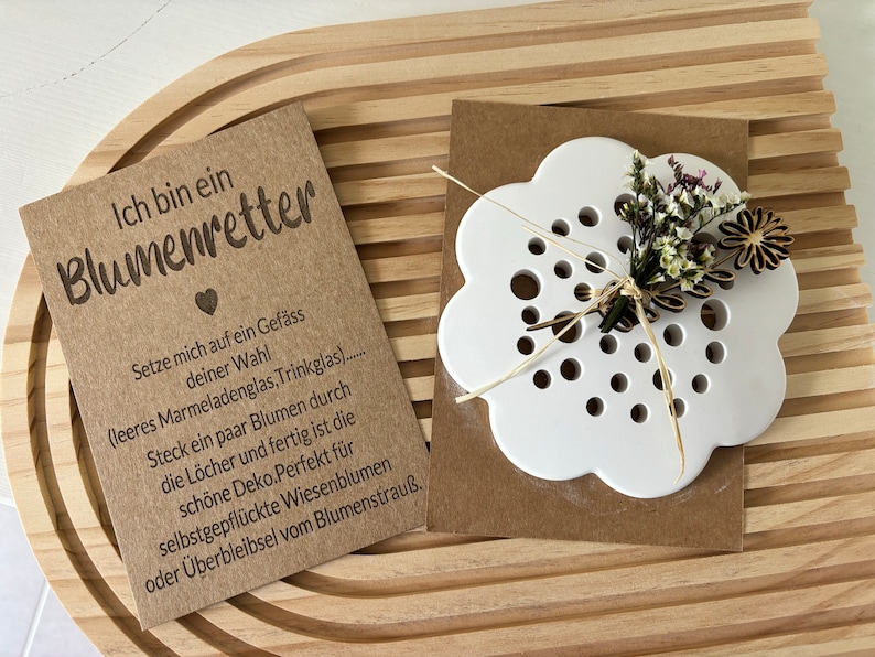 May include: A white ceramic flower-shaped vase with holes for stems. The vase is on a brown paper card with the text "Ich bin ein Blumenretter" and instructions for using the vase. The card and vase are on a wooden surface.