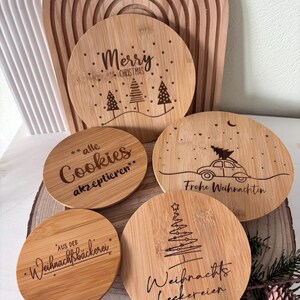 May include: A collection of round, wooden Christmas coasters with engraved designs and text. One coaster reads "Merry Christmas" with trees. Other coasters have phrases like "Cookies", "Frohe Weihnachten", and "Weihnachts Leckereien".