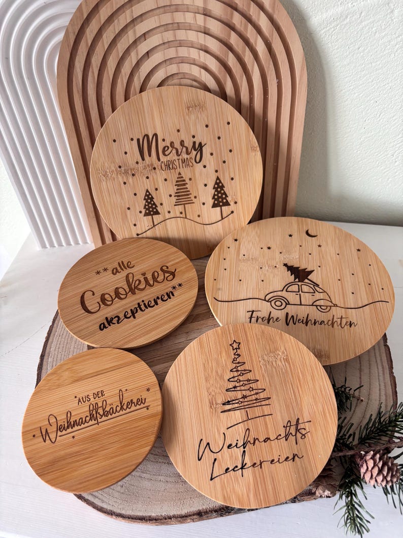 May include: A collection of round, wooden Christmas coasters with engraved designs and text. One coaster reads "Merry Christmas" with trees. Other coasters have phrases like "Cookies", "Frohe Weihnachten", and "Weihnachts Leckereien".