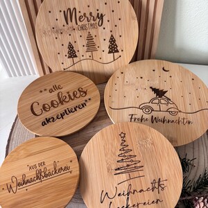 May include: A collection of round bamboo coasters with engraved Christmas-themed designs. One coaster reads "Merry Christmas" with trees, another says "Cookies akzeptieren", and others feature festive text and imagery. The coasters are arranged on a wooden surface.
