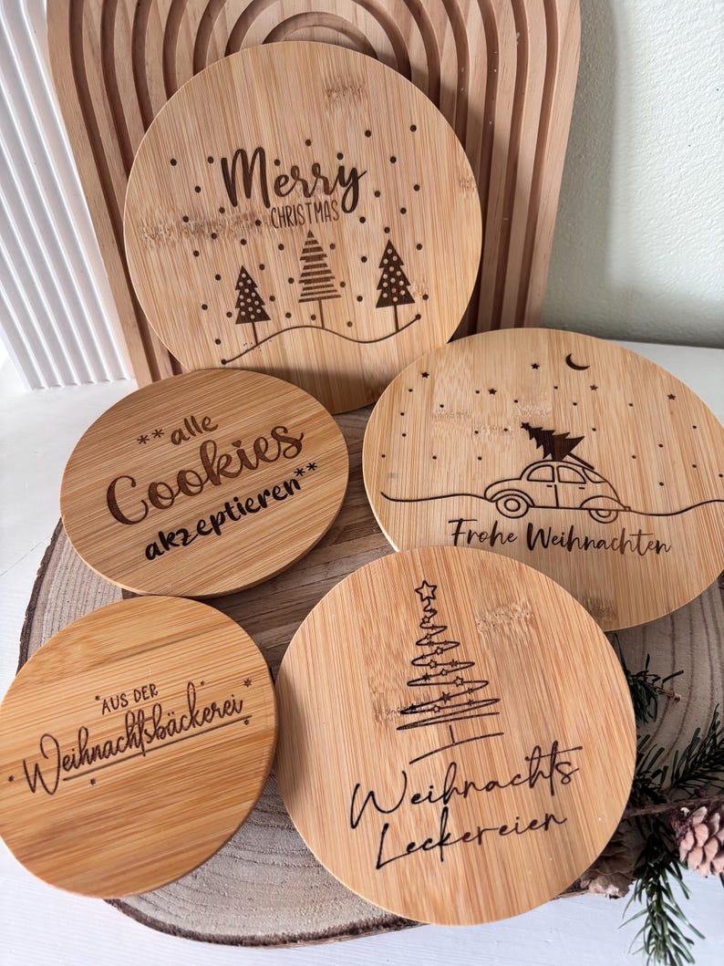 May include: A collection of round bamboo coasters with engraved Christmas-themed designs. One coaster reads "Merry Christmas" with trees, another says "Cookies akzeptieren", and others feature festive text and imagery. The coasters are arranged on a wooden surface.