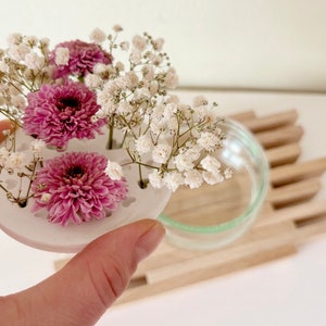 May include: A white ceramic flower frog with four holes for stems. The frog is holding pink and white flowers.