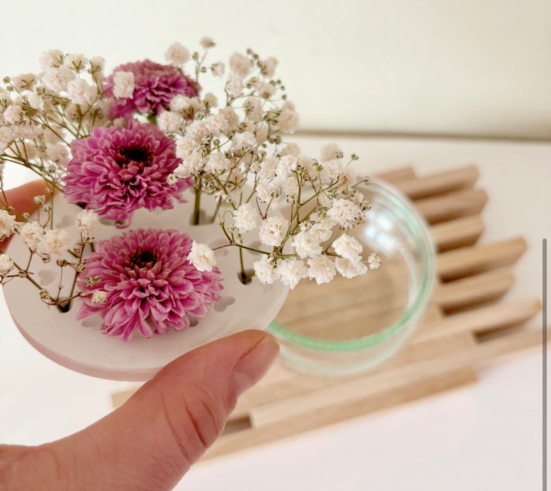 May include: A white ceramic flower frog with four holes for stems. The frog is holding pink and white flowers.