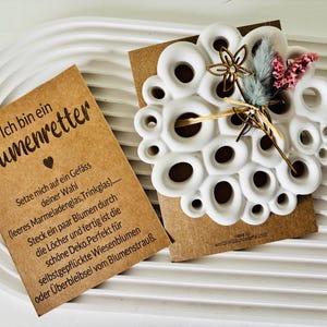 May include: A white ceramic flower frog with multiple holes, displayed on a brown card with German text. The frog is decorated with dried flowers and a gold-colored floral accent. The card reads "Ich bin ein Blumenretter."