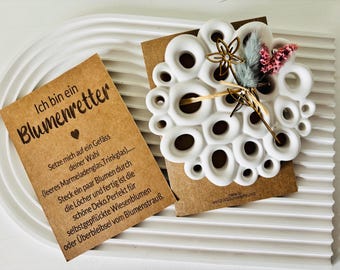 May include: A white ceramic flower frog with multiple holes, displayed on a brown card with German text. The frog is decorated with dried flowers and a gold-colored floral accent. The card reads "Ich bin ein Blumenretter."
