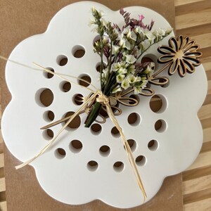 May include: White ceramic flower holder with a cloud-like shape and a pattern of small holes. A small bouquet of white and pink flowers is arranged in the holder, with a wooden flower cutout on the side.