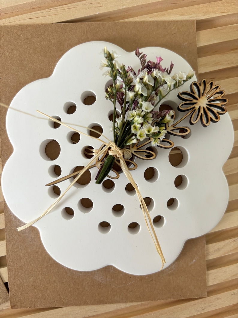May include: White ceramic flower holder with a cloud-like shape and a pattern of small holes. A small bouquet of white and pink flowers is arranged in the holder, with a wooden flower cutout on the side.