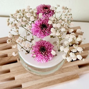 May include: A white ceramic vase with a decorative pattern of small circles and hearts. The vase is filled with pink and white flowers, including baby's breath and chrysanthemums. The vase is sitting on a wooden surface.