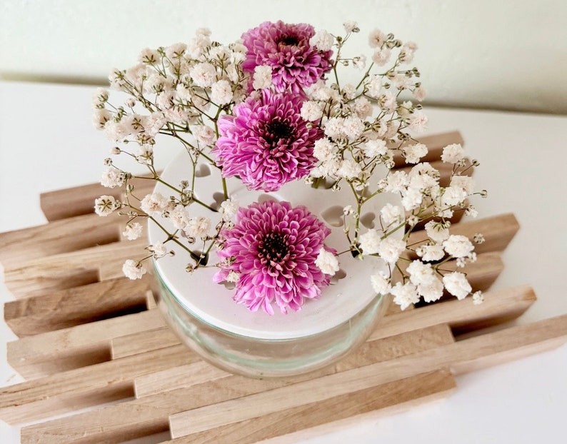 May include: A white ceramic vase with a decorative pattern of small circles and hearts. The vase is filled with pink and white flowers, including baby's breath and chrysanthemums. The vase is sitting on a wooden surface.