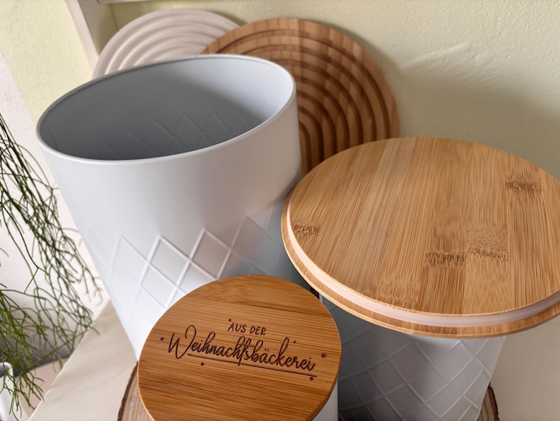 May include: A white metal container with a diamond pattern, paired with a wooden lid. The lid has a natural wood grain finish. A smaller wooden lid with text is also visible. The theme appears to be kitchen storage.