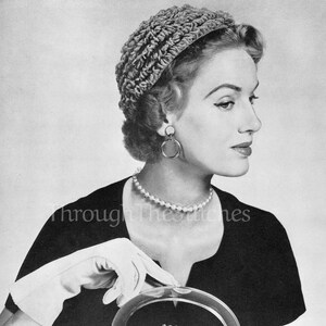 May include: A black and white photo of a woman wearing a crocheted hat and a black dress. She is holding a crocheted handbag with a clear plastic handle.