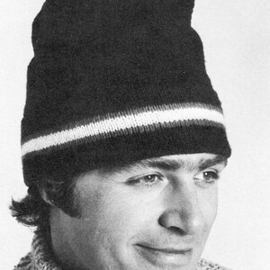 May include: A black knit hat with a white stripe near the brim. The hat is worn by a person wearing a textured, light-colored sweater. The image is in black and white.