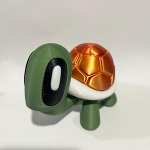 以下が含まれることがあります： 緑とオレンジの3Dプリントされた亀の置物で、白い甲羅をしています。亀は黒い目と白いハイライトを持っています。