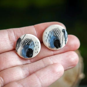 Peut inclure: Une paire de boucles d'oreilles rondes en céramique avec des motifs abstraits. Chaque boucle d'oreille présente une base blanche avec des coups de pinceau noirs, bleus et gris. Les boucles d'oreilles sont tenues dans une main ouverte.