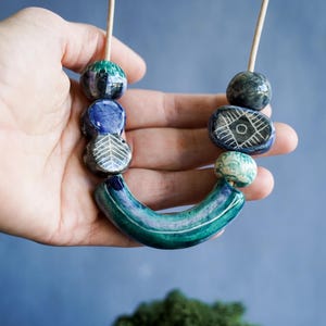May include: A necklace featuring a series of ceramic beads in shades of blue, green, and teal. The beads include round, oval, and curved shapes, with some displaying etched white geometric patterns like leaves and diamonds on their surfaces.