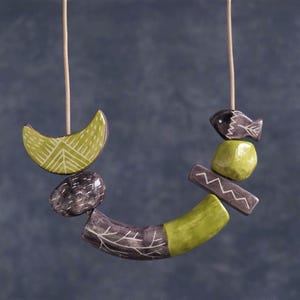 Puede incluir: Un collar único con una colección de cuentas de cerámica de varias formas y colores. El diseño incluye una media luna, un pez y formas geométricas en tonos verdes, grises y marrones, ensartadas en un cordón neutro.
