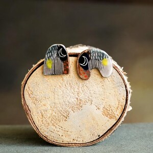 Puede incluir: Pendientes abstractos de plata con diseño de media luna. Los pendientes presentan una mezcla de colores, incluyendo negro, blanco, amarillo y rojo, dispuestos en un patrón geométrico. Los pendientes se exhiben sobre una rodaja de madera.