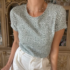 May include: A green and white patterned short-sleeved shirt with a white pearl necklace. The shirt is paired with white shorts.
