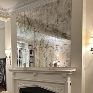 May include: A white fireplace mantel with a large antique mirror above it. The mirror is made up of 12 individual square tiles with a distressed, aged look. The mirror is reflecting the room behind it, including a bookshelf and a window with white blinds.