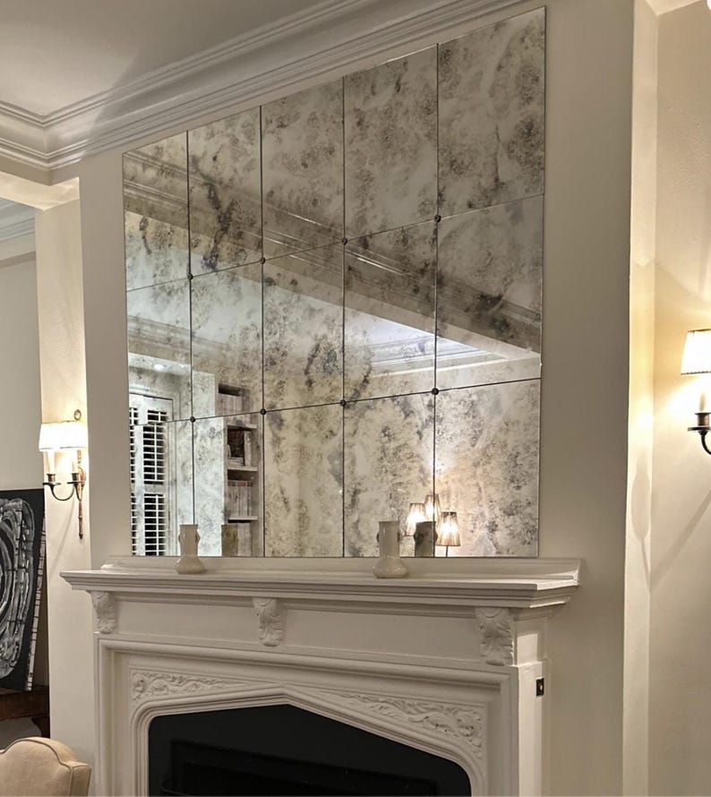 May include: A white fireplace mantel with a large antique mirror above it. The mirror is made up of 12 individual square tiles with a distressed, aged look. The mirror is reflecting the room behind it, including a bookshelf and a window with white blinds.
