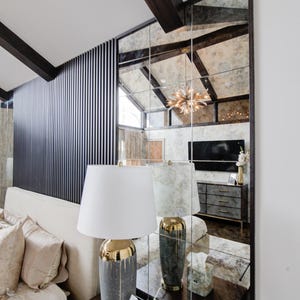 May include: A white lamp with a gold-trimmed base sits on a dark wood bedside table next to a white tissue box. The lamp is in front of a large mirrored wall with a black wood frame.
