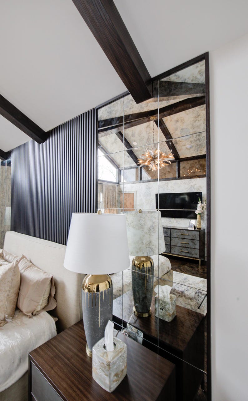 May include: A white lamp with a gold-trimmed base sits on a dark wood bedside table next to a white tissue box. The lamp is in front of a large mirrored wall with a black wood frame.
