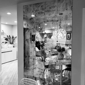 May include: A black and white photograph of a dining room featuring a large mirrored wall. The mirror is composed of numerous small squares and reflects a dining table with chairs. The table is laid with a vase of flowers and a chandelier hangs above it.