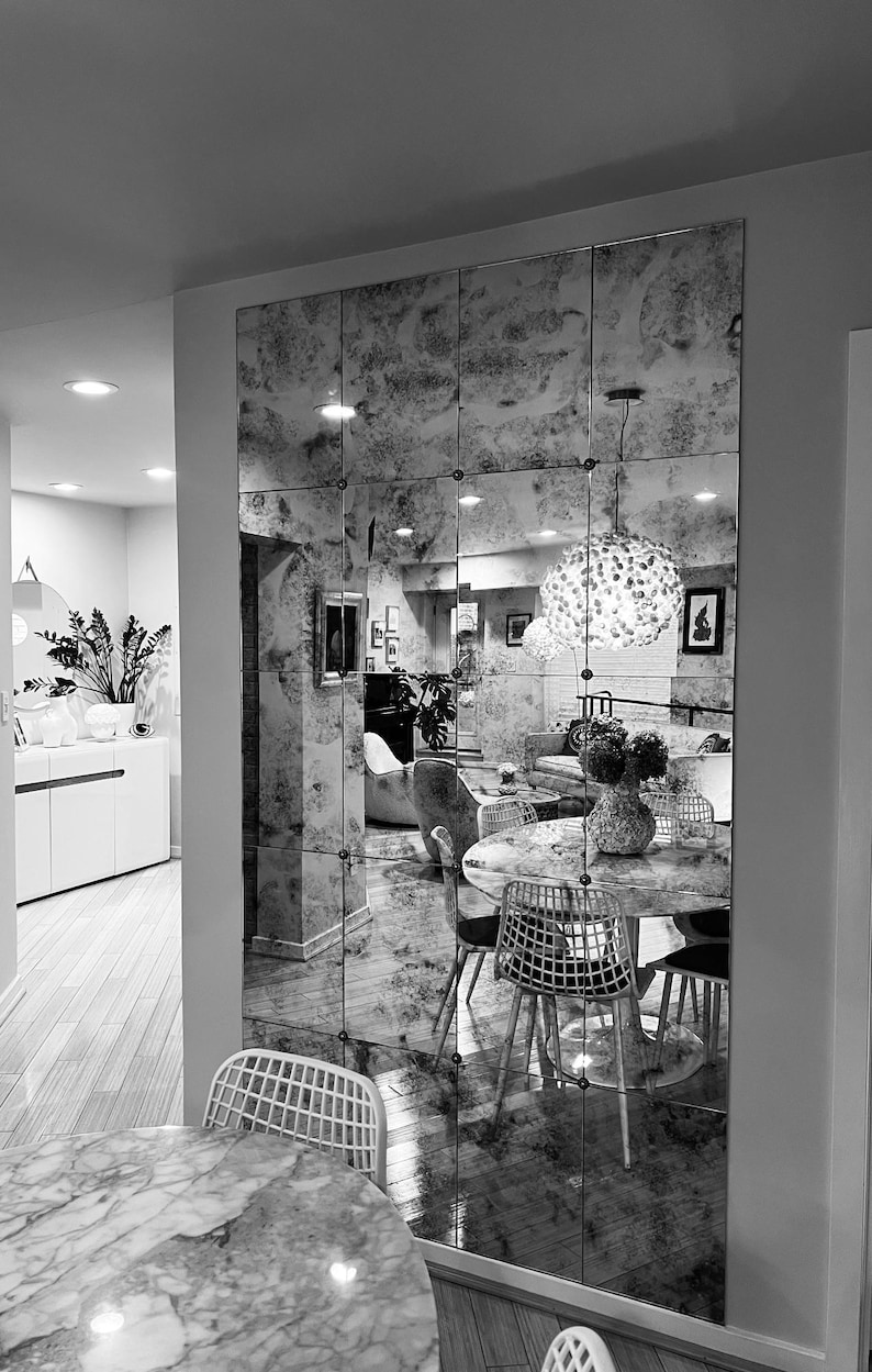 May include: A black and white photograph of a dining room featuring a large mirrored wall. The mirror is composed of numerous small squares and reflects a dining table with chairs. The table is laid with a vase of flowers and a chandelier hangs above it.