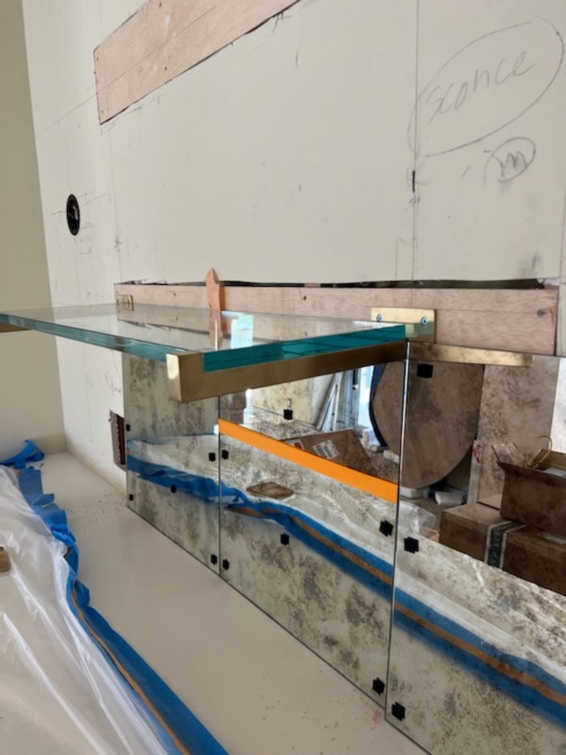 May include: A glass shelf with brass brackets is mounted to a wall with a mirrored backsplash. The shelf is angled and has a slight curve. The wall is unfinished and has a light brown wood panel.