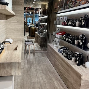 Puede incluir: Un interior de tienda minorista moderno con un suelo de baldosas grises, una pared de piedra gris y estantes llenos de varios productos, incluyendo botellas, velas y libros. Los estantes est&aacute;n iluminados con luces LED.