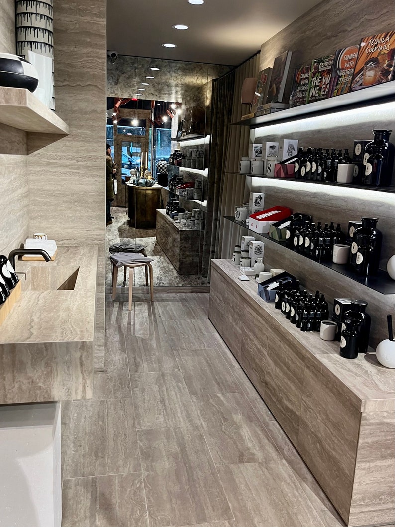 Puede incluir: Un interior de tienda minorista moderno con un suelo de baldosas grises, una pared de piedra gris y estantes llenos de varios productos, incluyendo botellas, velas y libros. Los estantes est&aacute;n iluminados con luces LED.