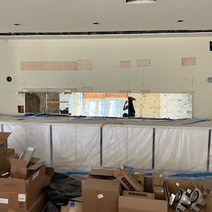 May include: A white countertop with a row of cabinets covered in plastic sheeting. The cabinets are in front of a wall with a mirror mosaic and several rectangular holes cut out. There are boxes and other construction materials in the foreground.