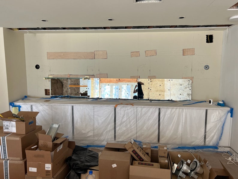 May include: A white countertop with a row of cabinets covered in plastic sheeting. The cabinets are in front of a wall with a mirror mosaic and several rectangular holes cut out. There are boxes and other construction materials in the foreground.