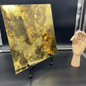 K&ouml;nnte beinhalten: Ein quadratisches St&uuml;ck goldfarbenes Glas mit einer marmorierten, strukturierten Oberfl&auml;che. Das Glas steht auf einem schwarzen St&auml;nder mit drei Beinen.