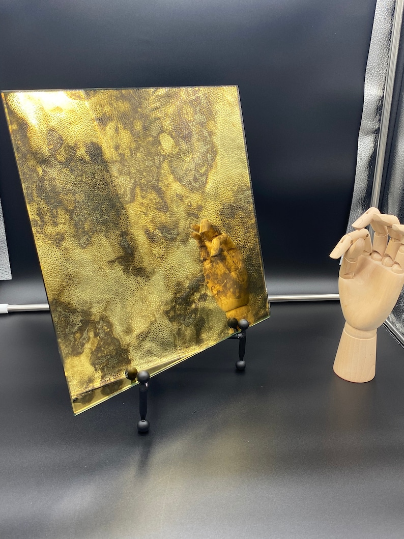 K&ouml;nnte beinhalten: Ein quadratisches St&uuml;ck goldfarbenes Glas mit einer marmorierten, strukturierten Oberfl&auml;che. Das Glas steht auf einem schwarzen St&auml;nder mit drei Beinen.