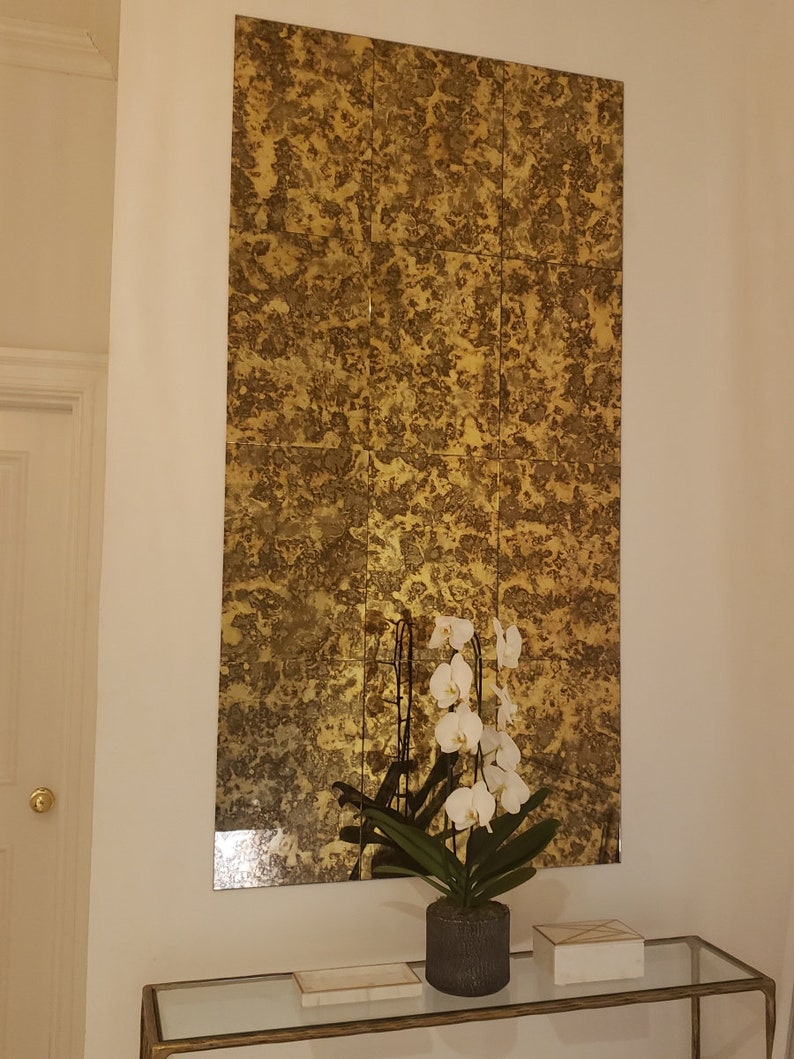 K&ouml;nnte beinhalten: Ein goldfarbener Spiegel mit einer abgenutzten Oberfl&auml;che ist an einer wei&szlig;en Wand &uuml;ber einem Glastisch montiert. Eine wei&szlig;e Orchideenpflanze in einem grauen Topf steht auf dem Tisch, zusammen mit zwei wei&szlig;en rechteckigen Boxen.