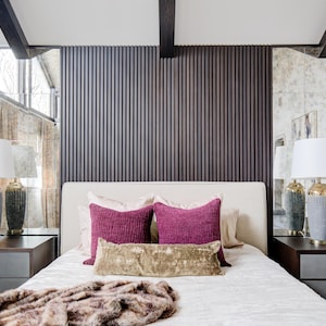 May include: A white bed with a white headboard and two pink throw pillows. The headboard is made of wood with vertical slats. There are two lamps on bedside tables on either side of the bed. The lamps have white shades and grey bases.