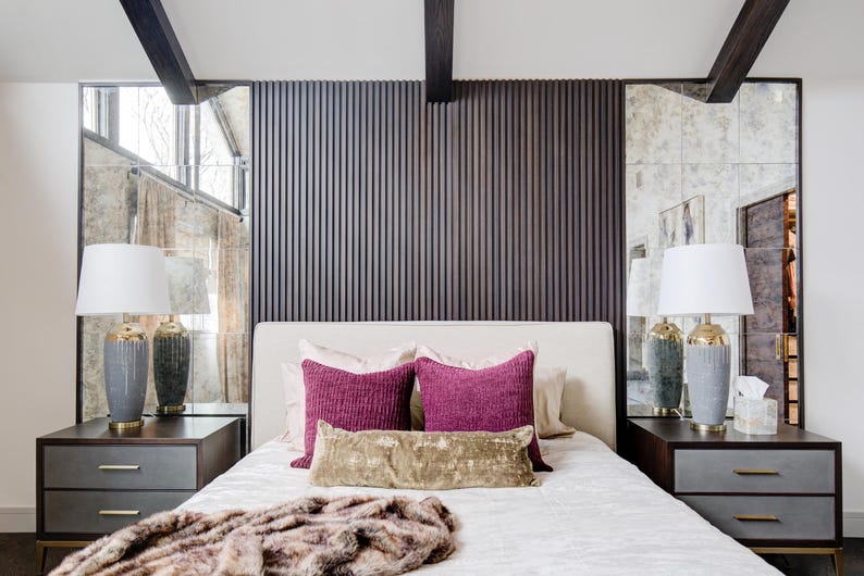 May include: A white bed with a white headboard and two pink throw pillows. The headboard is made of wood with vertical slats. There are two lamps on bedside tables on either side of the bed. The lamps have white shades and grey bases.