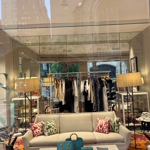 Puede incluir: Un sof&aacute; blanco con almohadas coloridas se encuentra en una tienda minorista. El sof&aacute; est&aacute; frente a una mesa de caf&eacute; blanca con dos bolsos de mano azules y dorados y una revista. La tienda tiene un estante de ropa en el fondo.