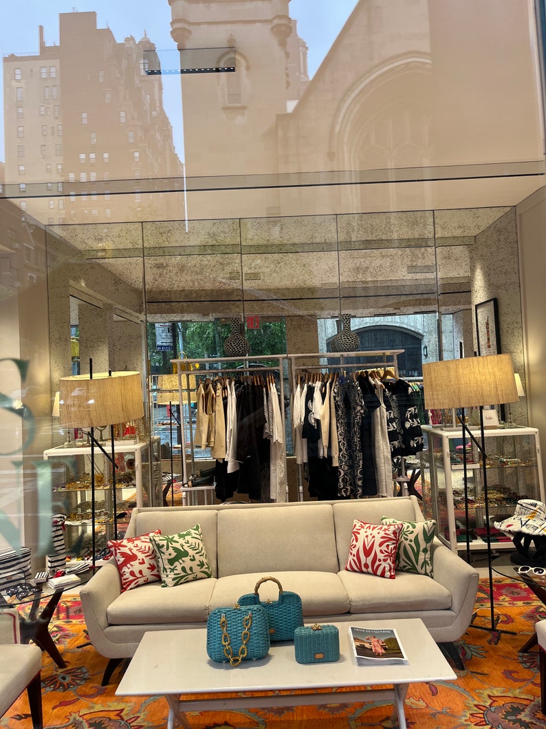 Puede incluir: Un sof&aacute; blanco con almohadas coloridas se encuentra en una tienda minorista. El sof&aacute; est&aacute; frente a una mesa de caf&eacute; blanca con dos bolsos de mano azules y dorados y una revista. La tienda tiene un estante de ropa en el fondo.