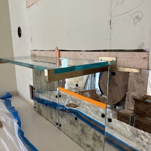 Puede incluir: Una estanter&iacute;a de vidrio con soportes de lat&oacute;n est&aacute; montada en una pared con un salpicadero de espejo. La estanter&iacute;a est&aacute; inclinada y tiene un tinte verde claro. El salpicadero de espejo tiene un aspecto desgastado y est&aacute; formado por varios paneles.
