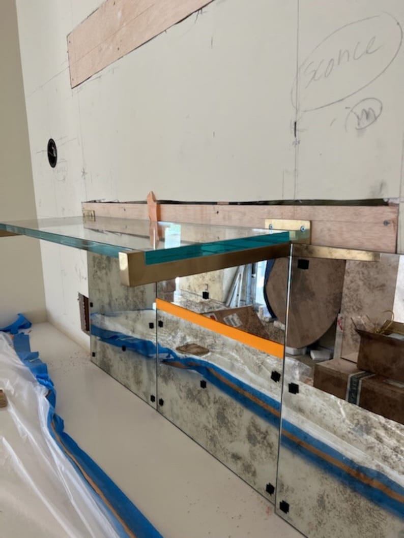 Puede incluir: Una estanter&iacute;a de vidrio con soportes de lat&oacute;n est&aacute; montada en una pared con un salpicadero de espejo. La estanter&iacute;a est&aacute; inclinada y tiene un tinte verde claro. El salpicadero de espejo tiene un aspecto desgastado y est&aacute; formado por varios paneles.