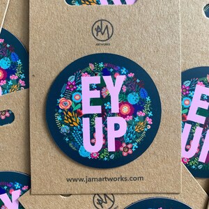 Può includere: Un adesivo rotondo con sfondo blu scuro e un design floreale rosa. Le parole "EY UP" sono scritte in lettere rosa al centro dell'adesivo.