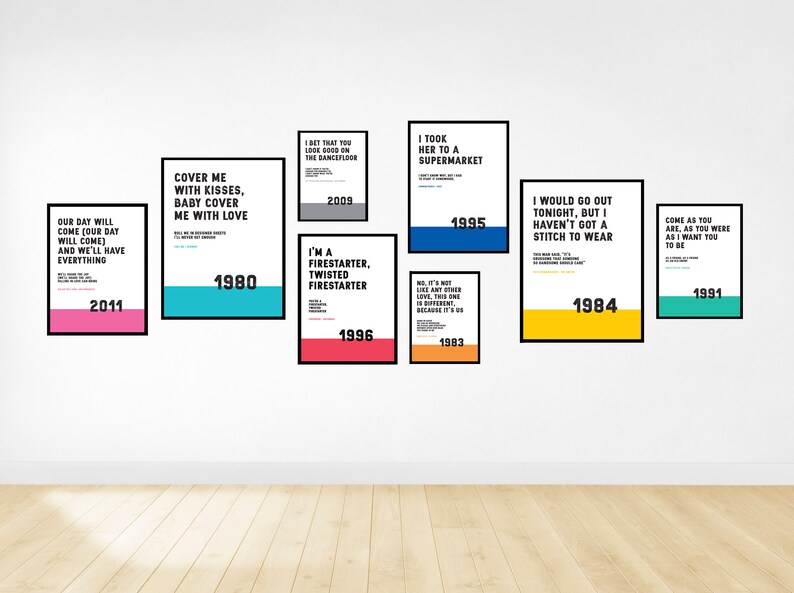 Puede incluir: Una pared blanca con nueve carteles enmarcados, cada uno con una letra de canci&oacute;n diferente y el a&ntilde;o en que se public&oacute;. Los carteles son de varios colores, incluyendo rosa, azul, amarillo y verde.